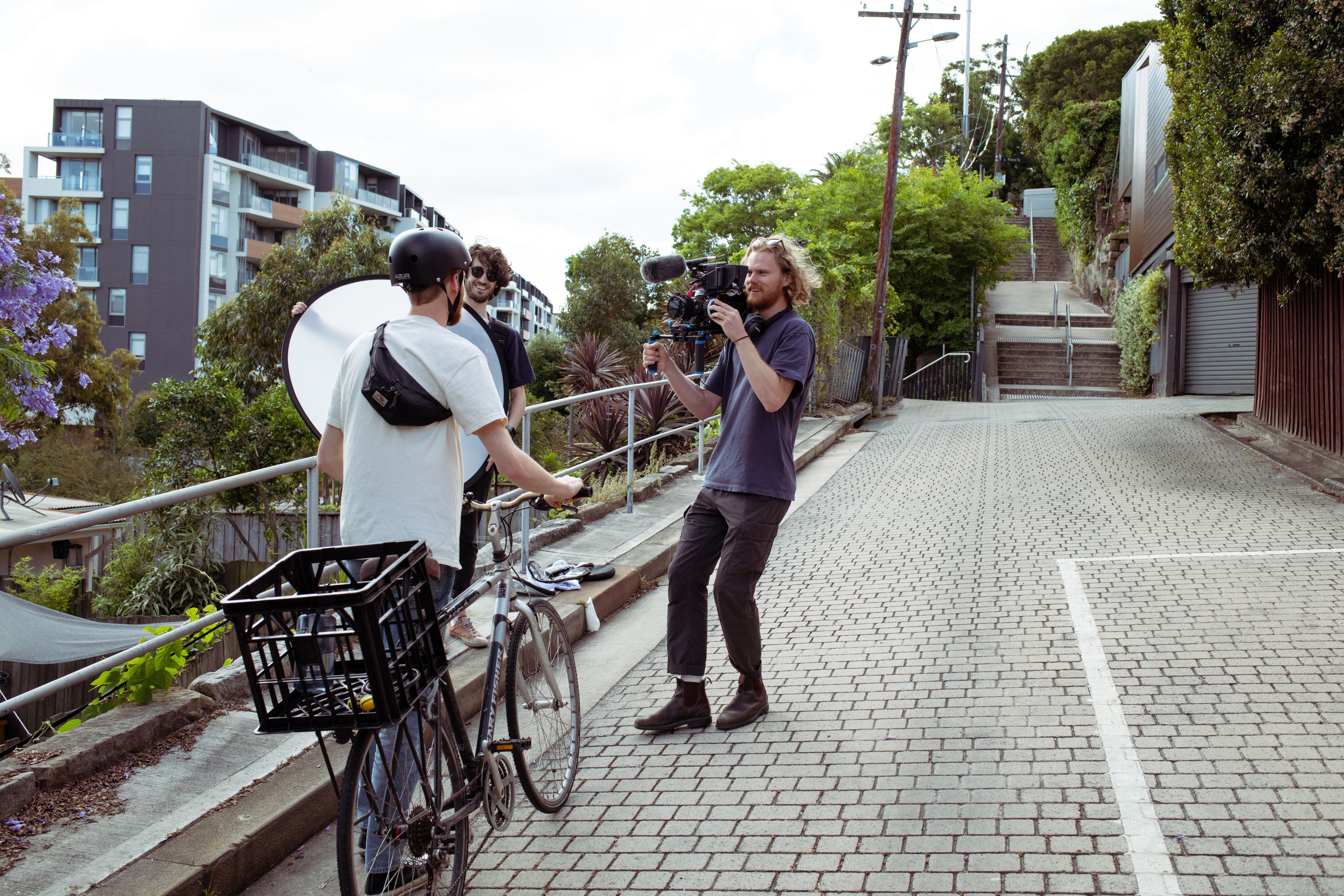 A person on a bicycle interacts with a solopreneur filmmaker holding a camera, while another person holds a reflector.