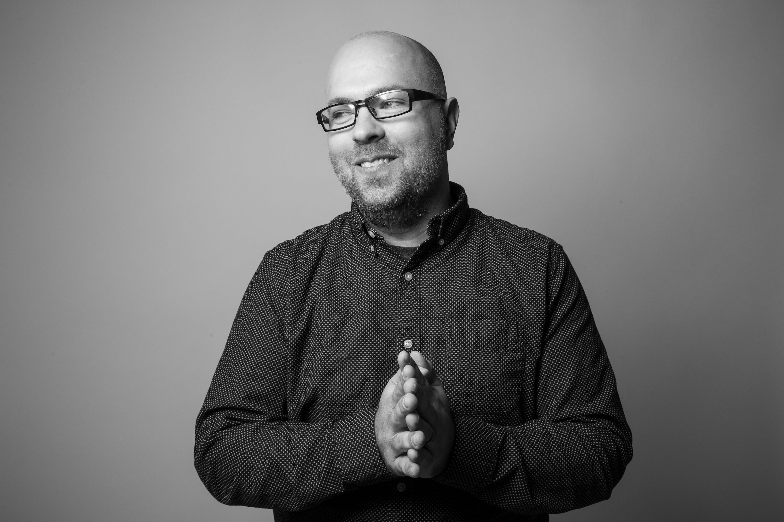 Adam Nielson wearing a black button down and glasses standing for a black and white photo