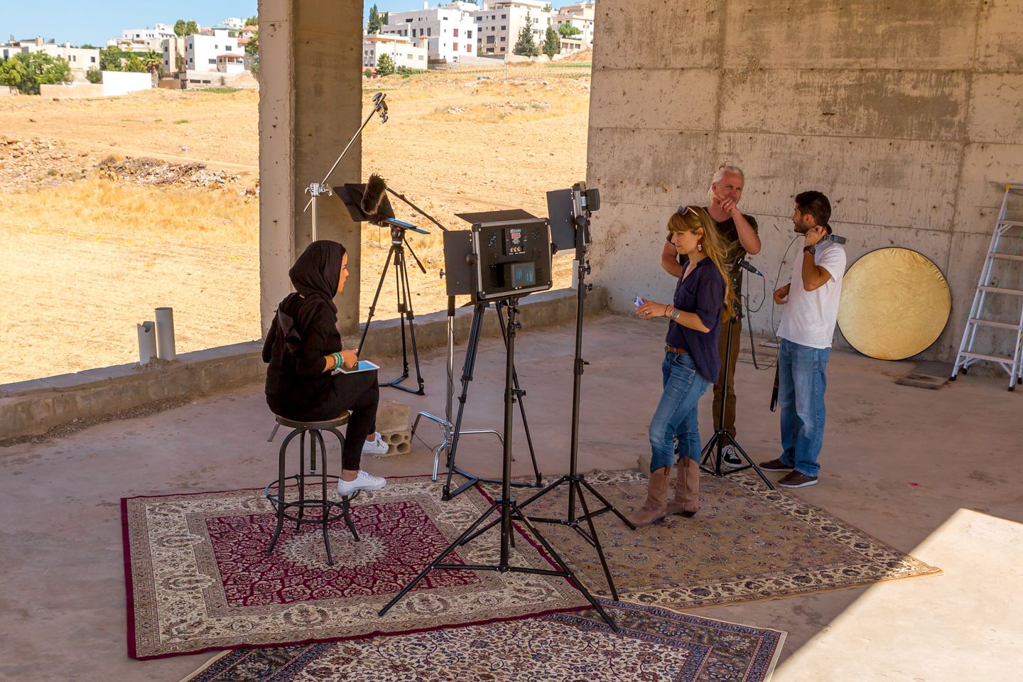 One person sitting on a stool infront of a camera set up with three individuals behind the camera and lights.