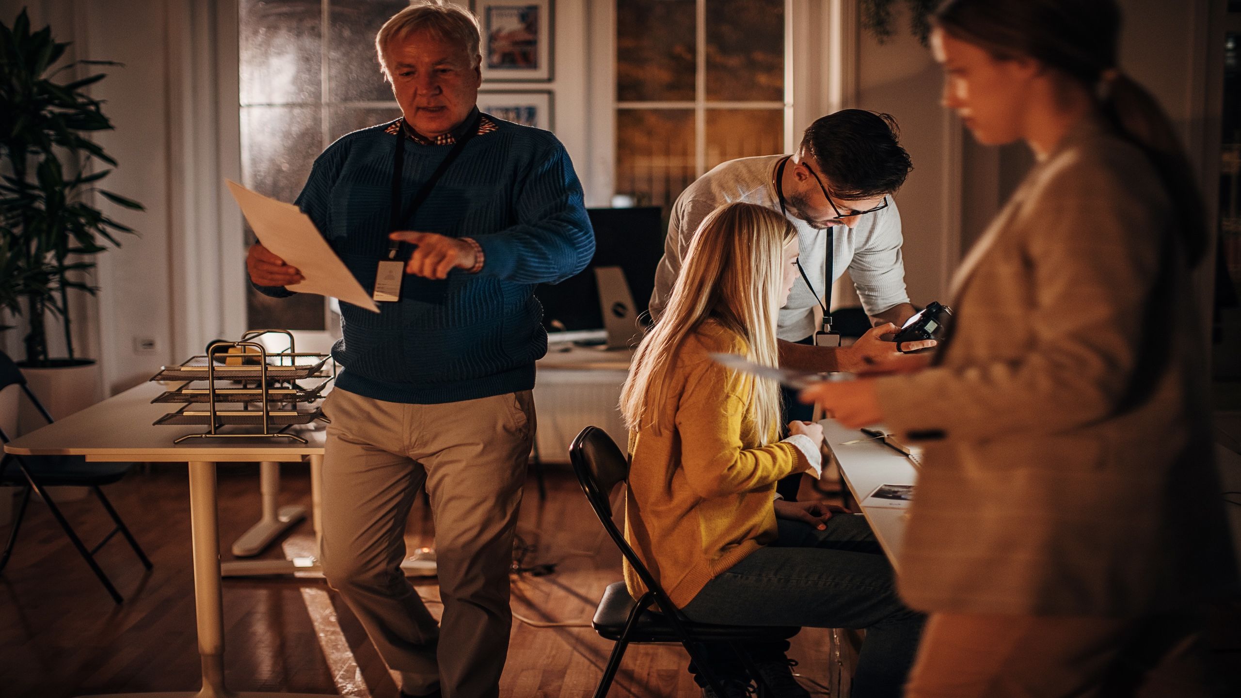 Four people in a dim room, two working at a desk together and two looking at printed stories for review. 