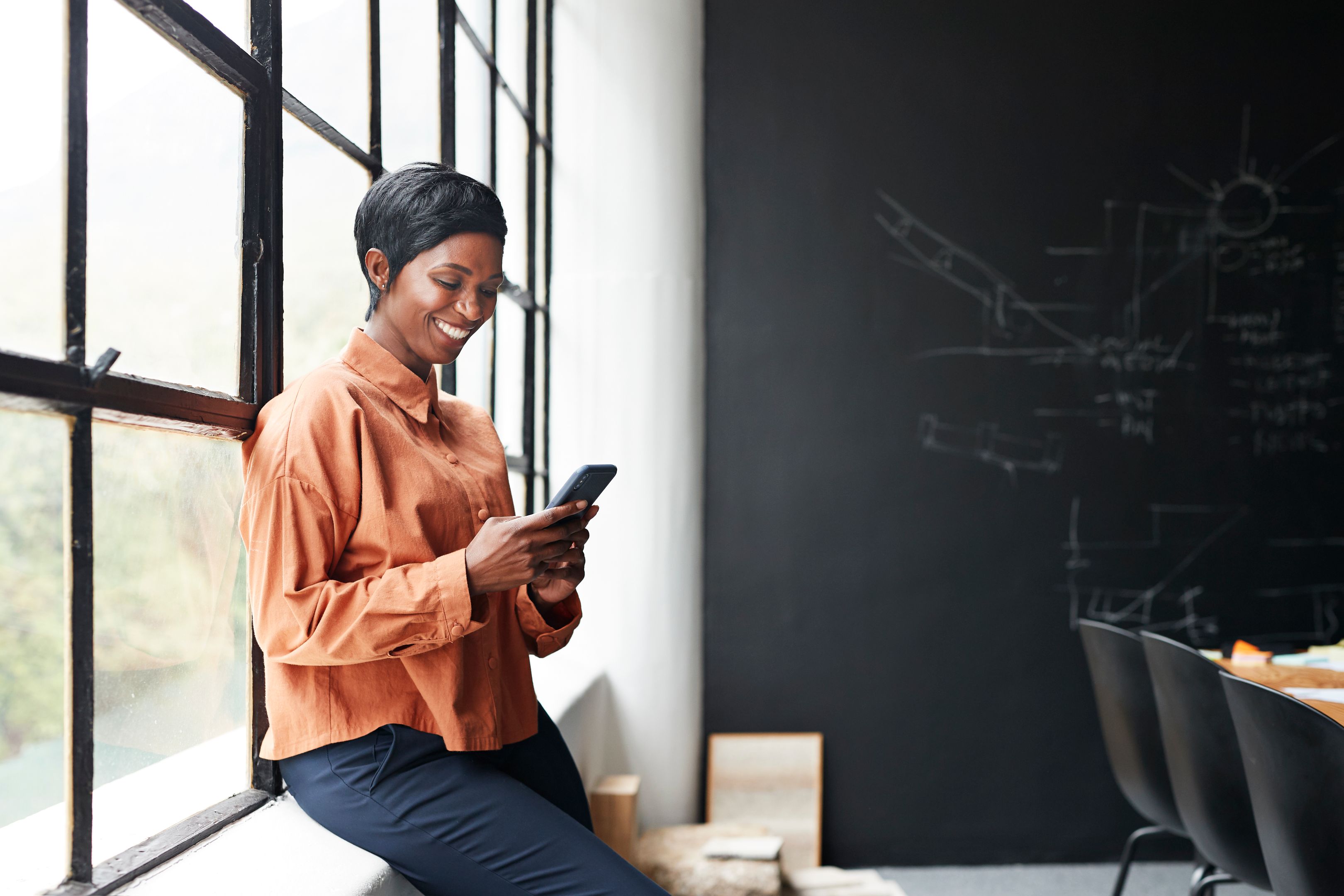 A person sitting on a windowsill smiling at their mobile device.