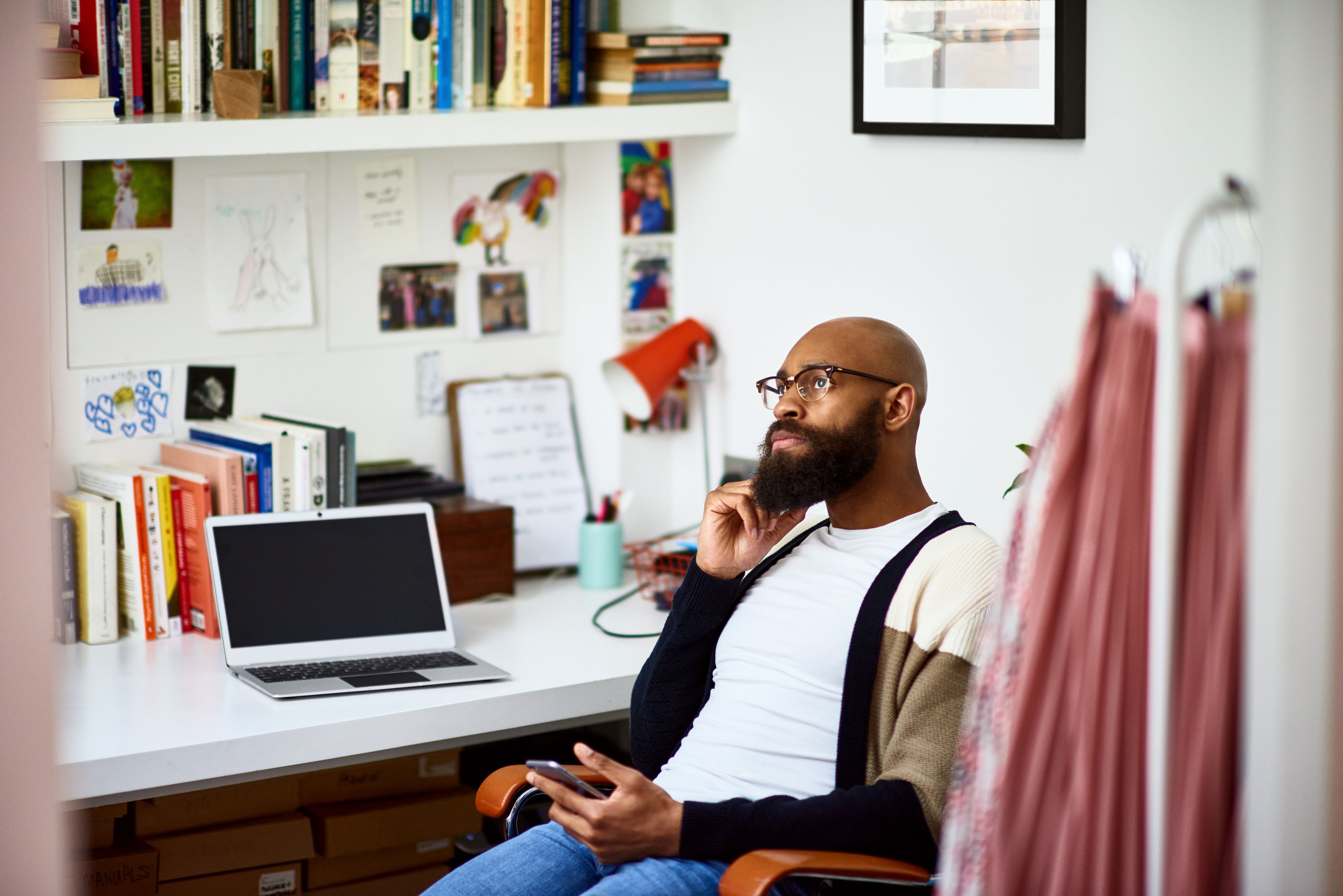 A person sits in a chair at a desk, stroking their beard with one hand, while the other holds a mobile device on their lap.