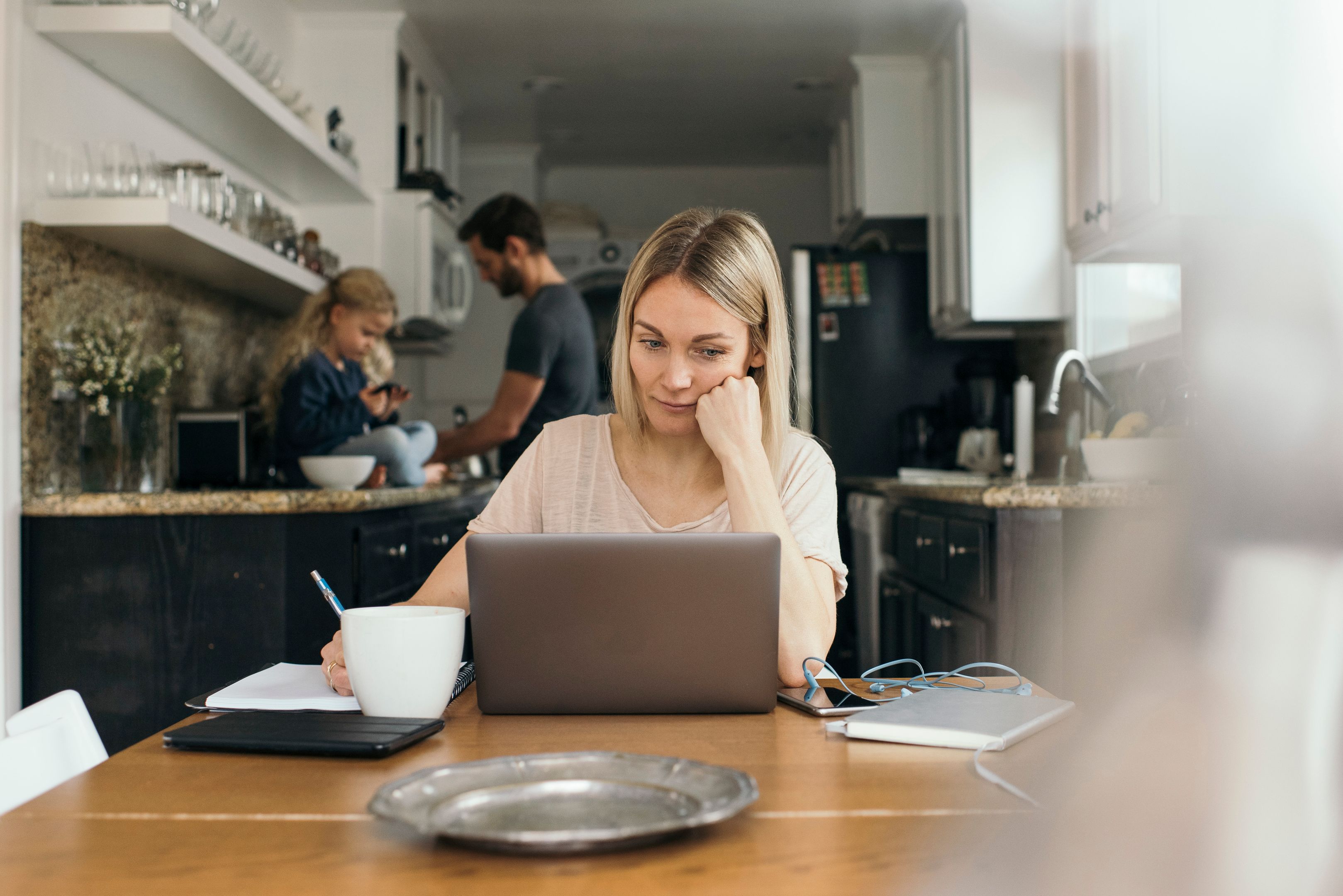 Parent works on a laptop at a kitchen table, child sits on the counter with an adult nearby.