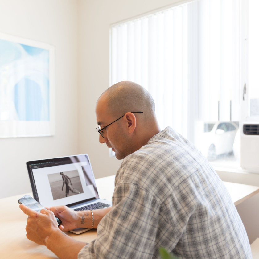 A photographer looks at their mobile device. A photo for a client is displayed on a laptop screen in the background.