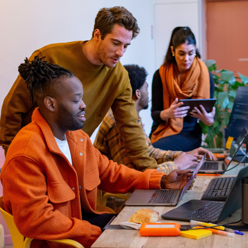 Quattro persone, due sedute e due in piedi, collaborano a un progetto utilizzando laptop e tablet.